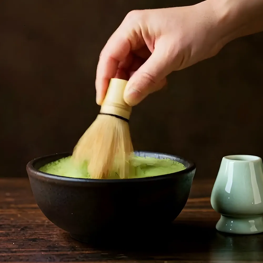 A hand whisking matcha to a foam with the bamboo whisk, the holder beside the bowl